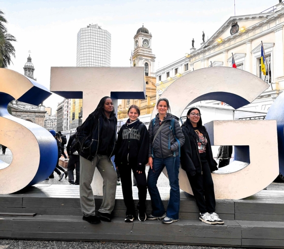 Three female students with a female professor in front of sign that reads STG