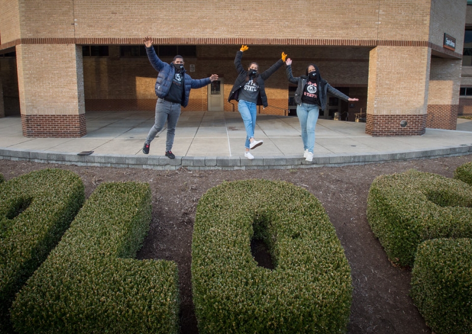 Three Buffalo State students wearing masks stand over the Commencement garden in front of Sports Arena