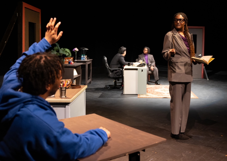 Student sitting at desk with hand raised, teacher pointing at and addressing him