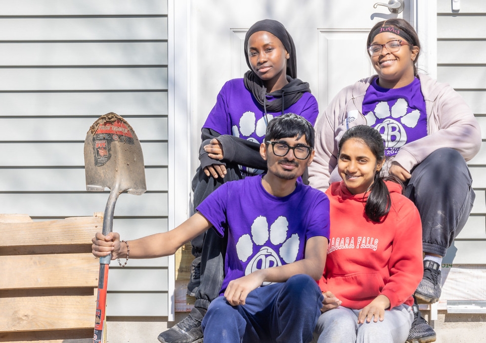 Four smiling Buffalo State students seated on steps, one holding shovel